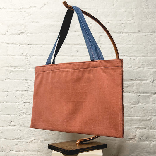 Pink Corduroy bag, with blue stitching on the bag proper and pink stitching on the blue strap. Hangs on a bent copper stand with a white brick background.